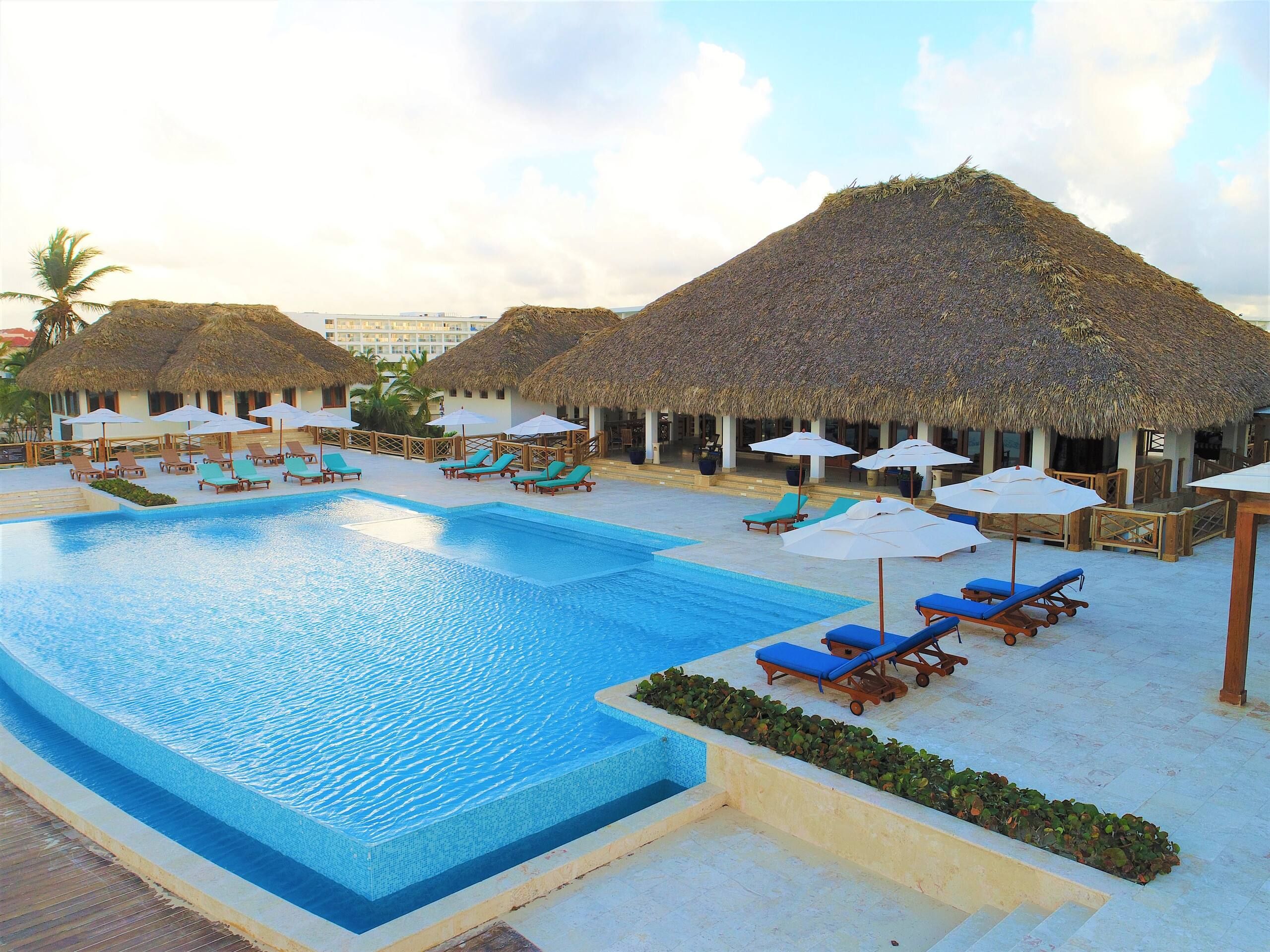Piscina del Cana Bay Beach Club con palapas de techo de paja, sombrillas y tumbonas azules