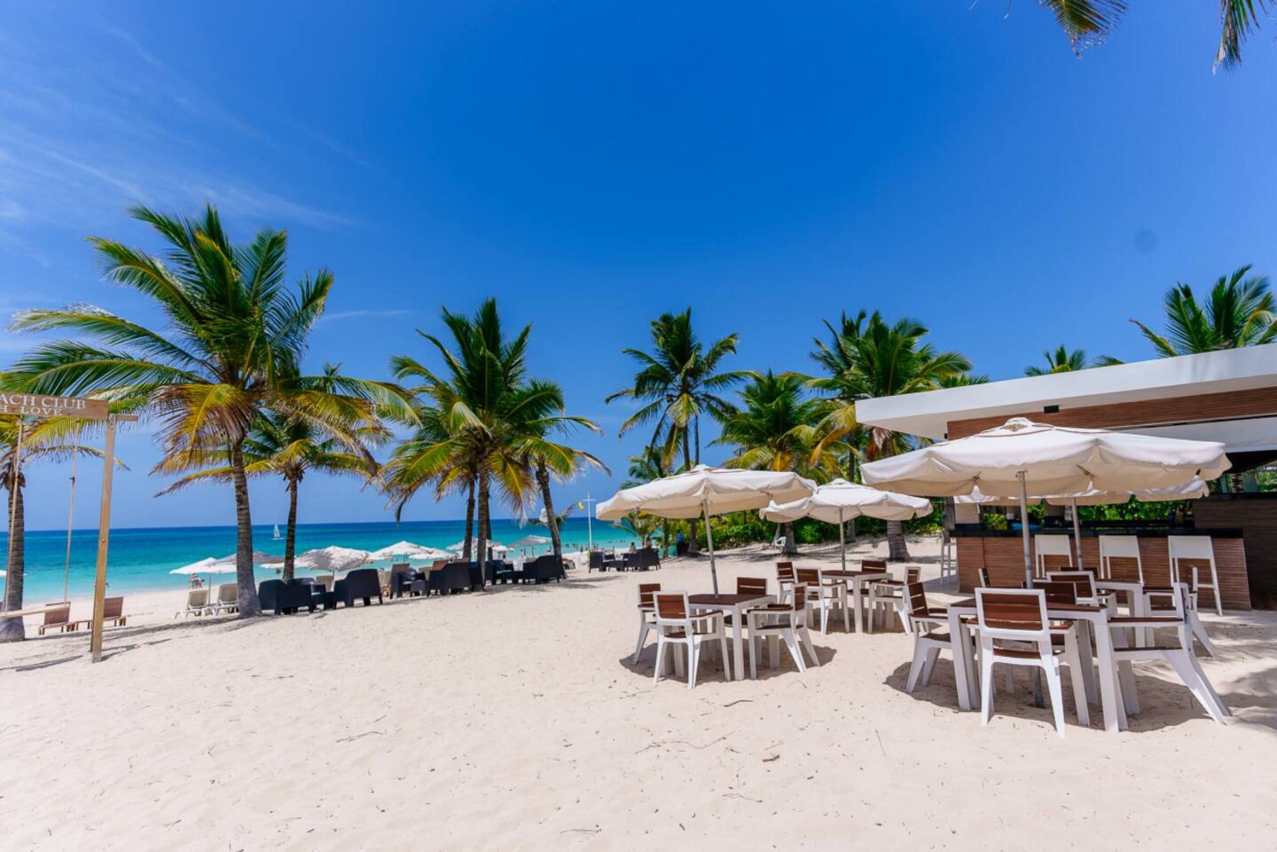 Playa de arena blanca con palmeras, sombrillas y mesas en el beach club